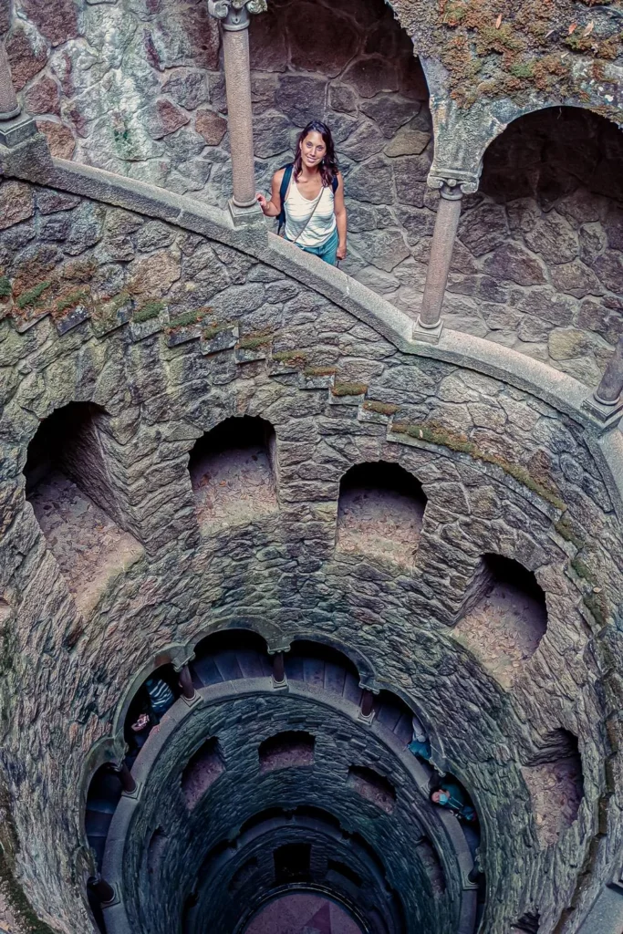 Caroline stands on the spiral staircase of the initiatory well at Quinta da Regaleira in Sintra, surrounded by stone walls covered in moss.
