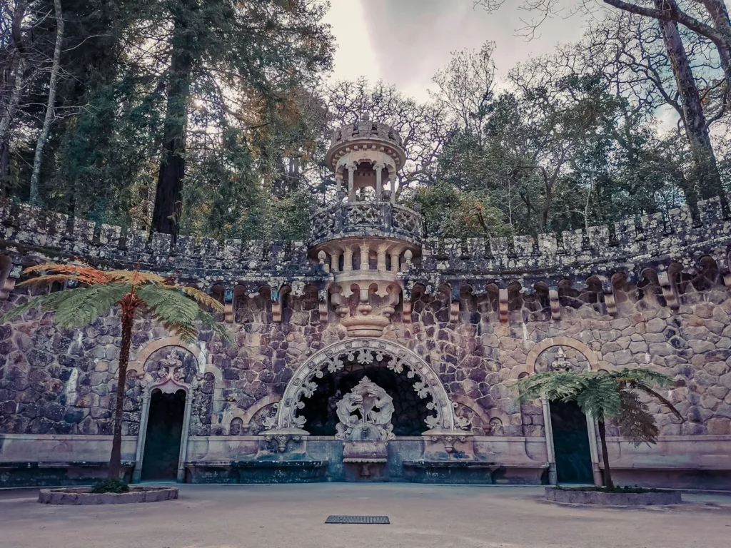 Ornate stone facade and sculpted fountain in the gardens of Quinta da Regaleira in Sintra, surrounded by vegetation and trees in March 2022.