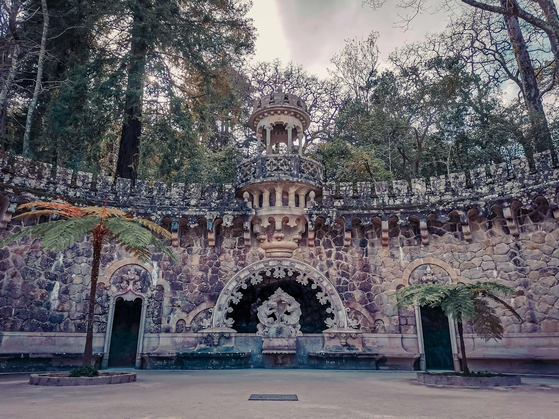 Ornate stone facade and sculpted fountain in the gardens of Quinta da Regaleira in Sintra, surrounded by vegetation and trees in March 2022.