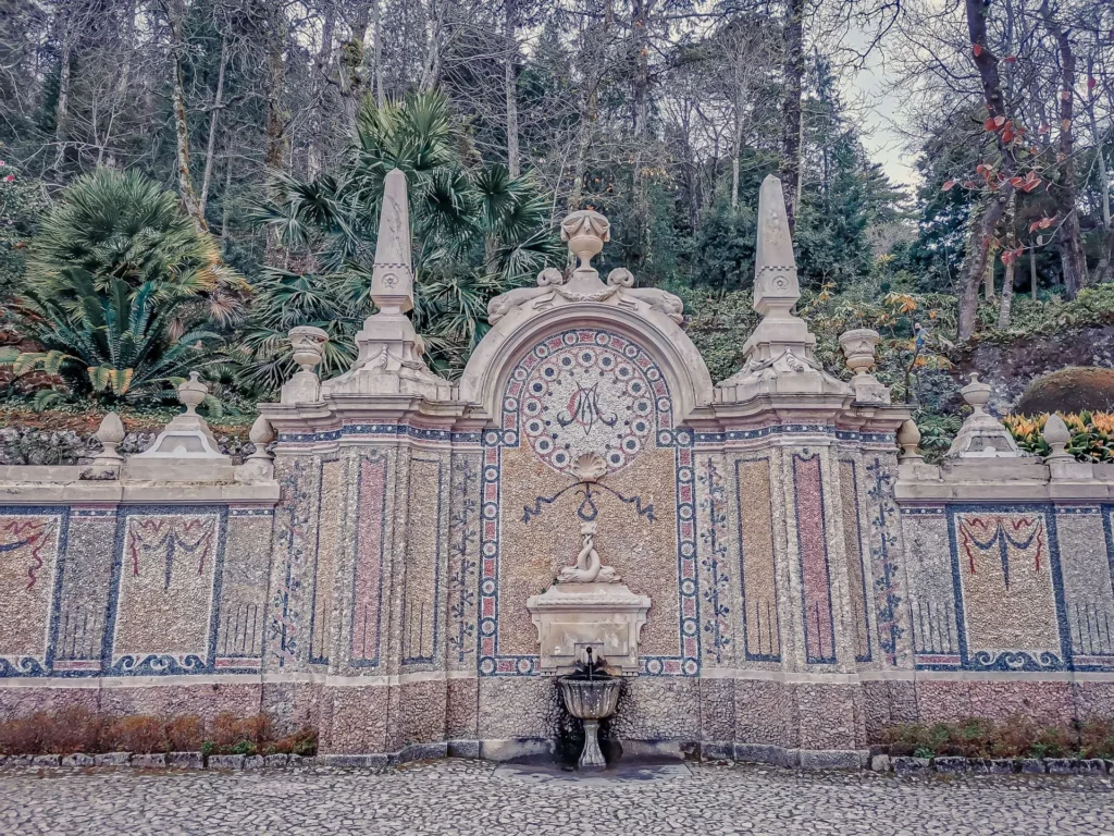 Ornamental stone fountain decorated with colorful mosaics in the gardens of Quinta da Regaleira, surrounded by dense vegetation in March 2022.