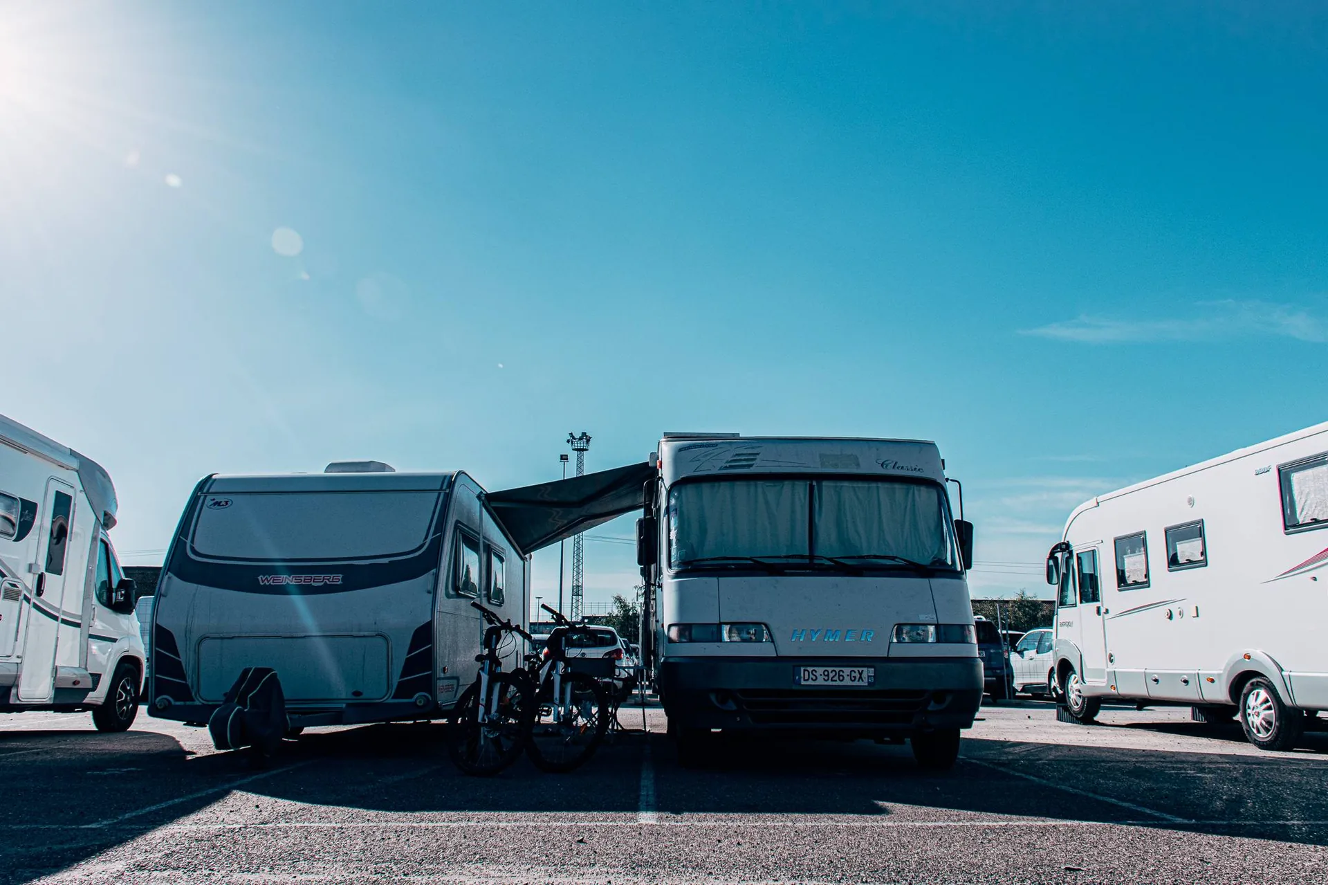 Alignement de camping-cars et caravanes sur un parking ensoleillé à Séville, Andalousie, avec deux vélos stationnés devant. Ambiance de voyage itinérant sous un ciel bleu.