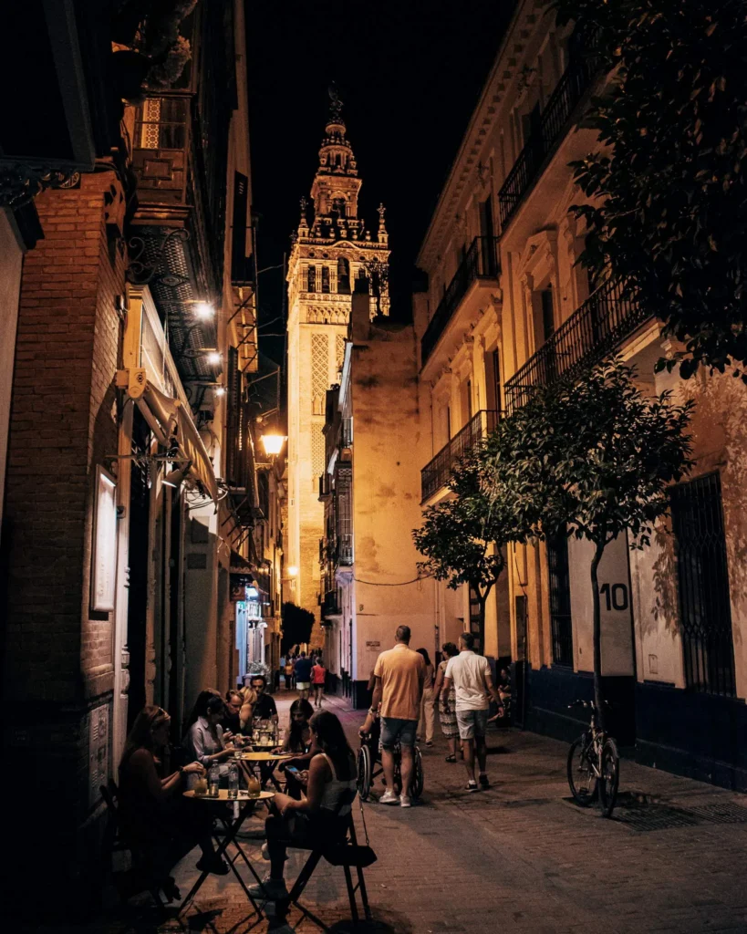 Rue animée de Séville en soirée, avec des personnes attablées en terrasse et la Giralda illuminée en arrière-plan.
