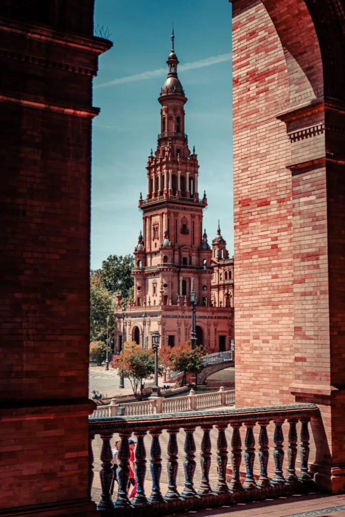 Vue sur la tour emblématique de la Plaza de España à Séville, prise depuis une arcade en brique sous un ciel dégagé.