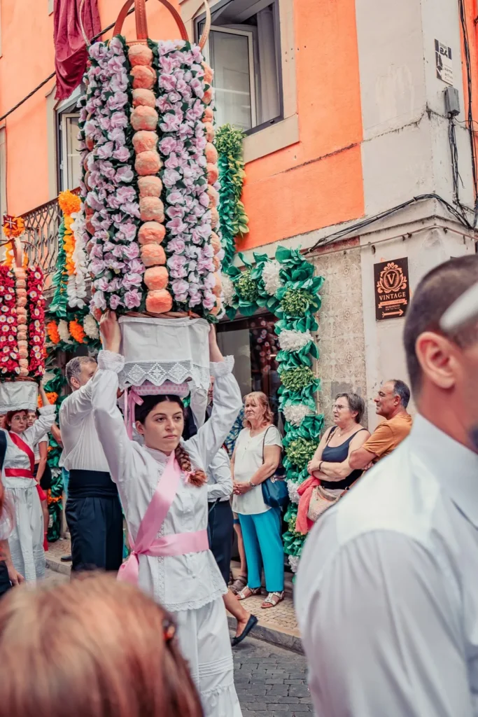 Femme porte un tabuleiro décoré de fleurs lors d'un défilé traditionnel dans une rue animée de Tomar pendant le Festival des Tabuleiros, sous le regard de spectateurs.