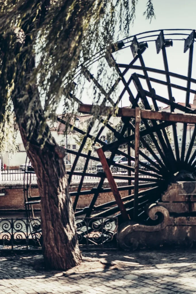 Roue à eau ancienne en bois au bord d'une rivière dans le centre historique de Tomar, sous un arbre, avec des bâtiments traditionnels en arrière-plan.