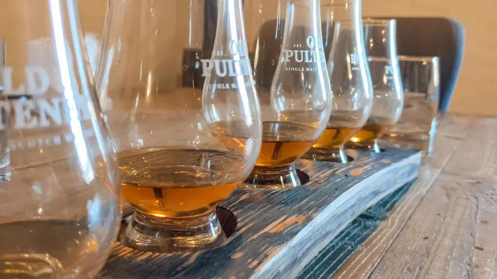 Old Pulteney whisky tasting with glasses lined up on a wooden table at the Wick distillery in Scotland, summer.