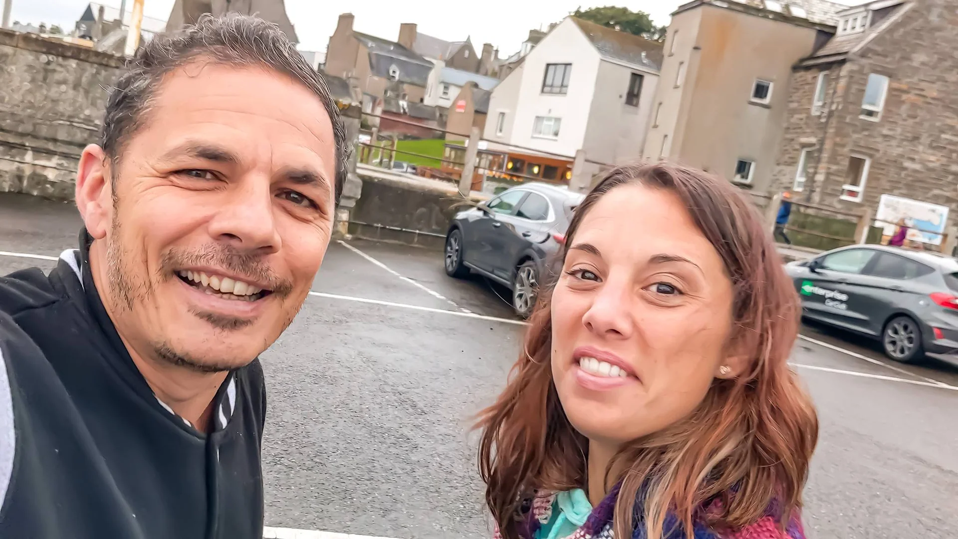 Xavier and Caroline smiling in a parking lot surrounded by typical stone buildings in Wick