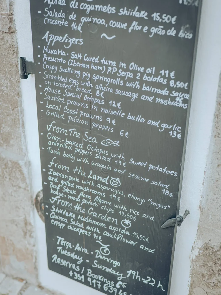 Menu board displayed at the entrance of the Case de Pasto restaurant in Tavira, Algarve, offering Portuguese dishes made from local products, handwritten in English and Portuguese.