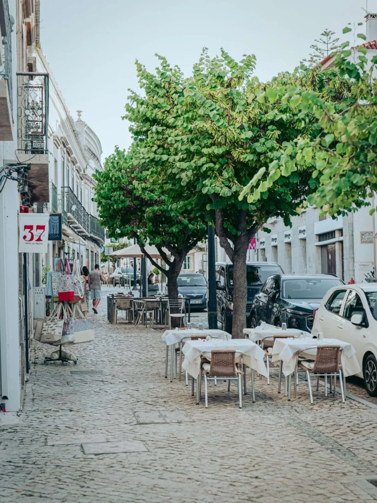 Terrace of the restaurant Case de Pasto 37 in Tavira, Algarve, with tables set up on a cobbled street lined with trees and typical buildings.
