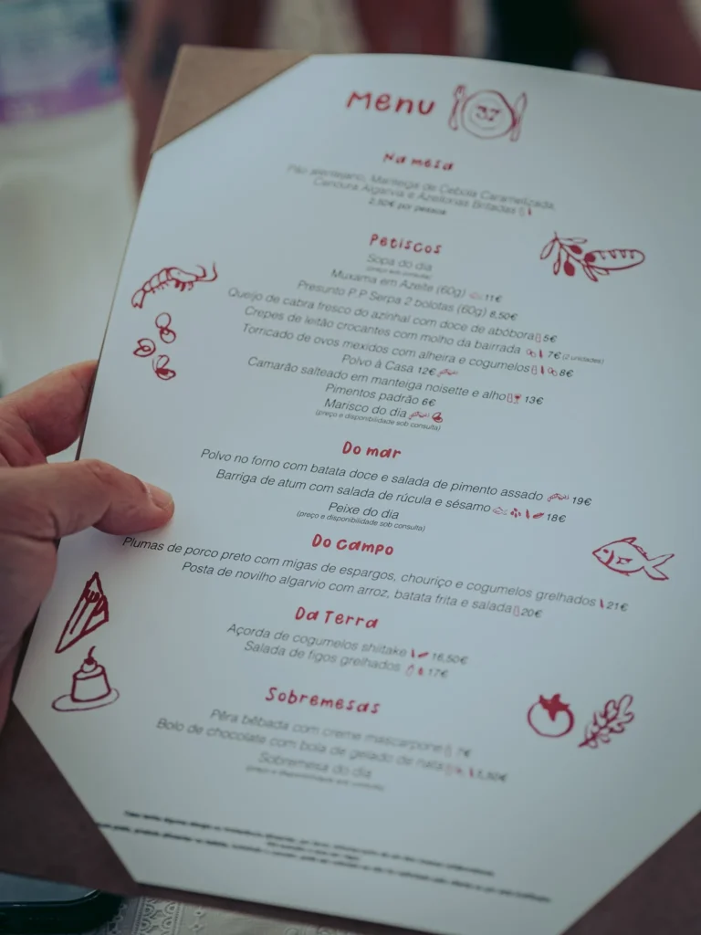 Menu of the Case de Pasto restaurant in Tavira, Algarve, displayed on a table, with a hand holding the menu. The dishes offered highlight Portuguese specialties and local products.
