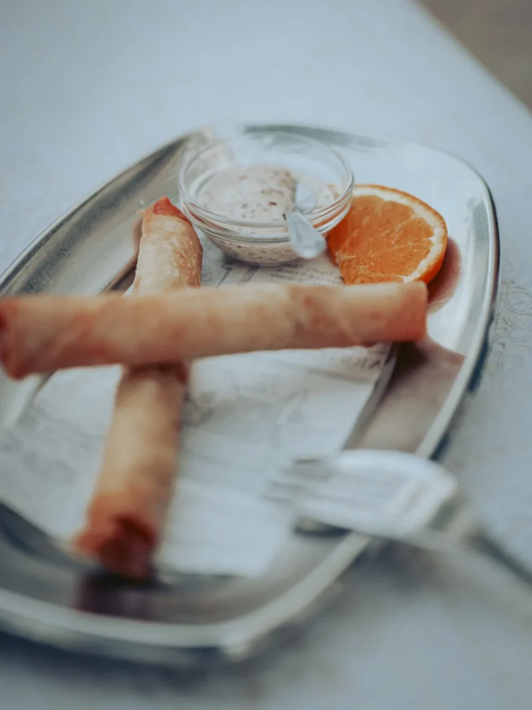 Metal plate containing two crispy leitão pancakes accompanied by a sauce in a small glass and a slice of orange, on a set table at the Case de Pasto restaurant in Tavira, Algarve.