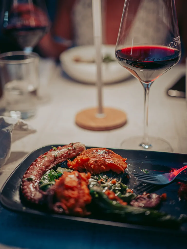 Plate of oven-baked octopus with sweet potato and roasted pepper salad on a restaurant table in Tavira, Algarve, with a glass of red wine beside it.