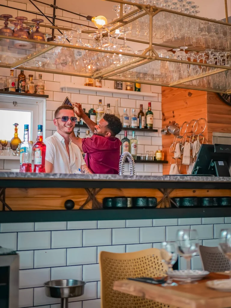 Bruno und Chris hinter der Theke eines Restaurants in Tavira, warme Atmosphäre mit Alkoholflaschen und hängenden Gläsern. Modernes Dekor mit weißen Fliesen und Holz.