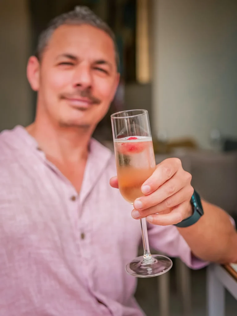 Xavier sitzt an einem Tisch im Restaurant Coisa Linda in Tavira, hebt ein Glas prickelnden Cocktails mit einer Himbeere, sommerliche und entspannte Atmosphäre.