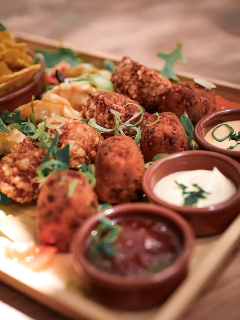 Sortiment von Spezialitäten auf einem Holzbrett im Restaurant Coisa Linda in Tavira, einschließlich indischer Piadjules, brasilianischer Tapioca-Dadines, Schweinegyozas und Hummus mit getrockneten Tomaten, begleitet von Saucen.