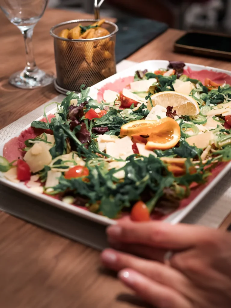 Ein Rindfleisch-Carpaccio, garni mit Rucola, Kirschtomaten, Käseflocken und Zitrusfrüchten, wird auf einem Holztisch in einem Restaurant in Tavira, Algarve, serviert. Ein kleiner Korb mit Pommes Frites begleitet das Gericht.