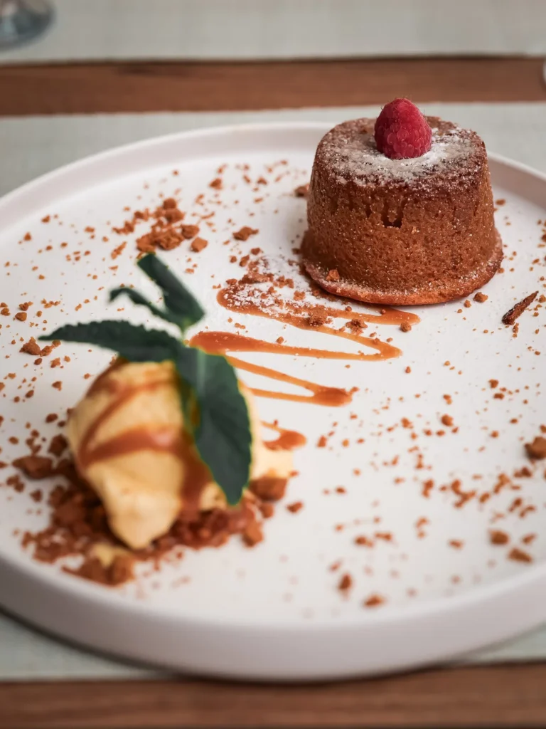Dessert Bolo de Leite, serviert auf einem weißen Teller, begleitet von einer Kugel Eis und dekoriert mit einer Himbeere und Karamell, im Restaurant Coisa Linda in Tavira.