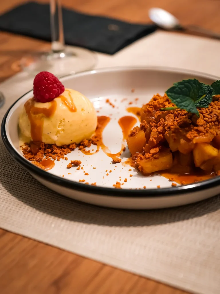 Crumble mit Äpfeln, serviert mit einer Kugel Vanilleeis und einer Himbeere, auf einem gedeckten Tisch im Restaurant Coisa Linda in Tavira.