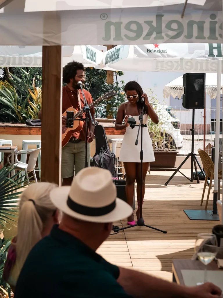 Duo von Musikern, die auf der Terrasse im Restaurant Coisa Linda in Tavira unter Heineken-Sonnenschirmen spielen, vor einem Publikum an einem sommerlichen Tag.