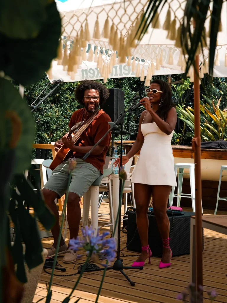 Sängerin singt am Mikrofon, begleitet von einem Gitarristen auf der sonnigen Terrasse eines Restaurants in Tavira, Algarve, Portugal, unter dekorativen Sonnenschirmen.