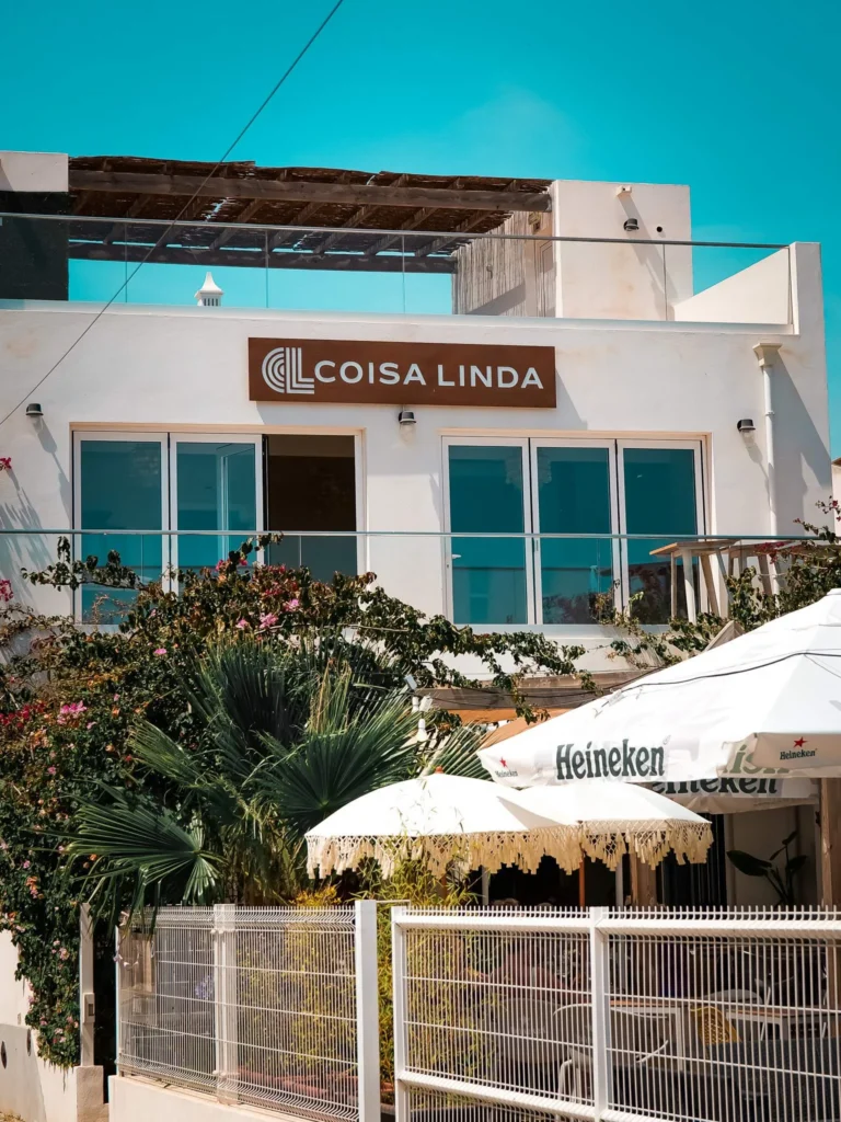 Fassade des Restaurants Coisa Linda in Tavira, Algarve, mit schattiger Terrasse unter Sonnenschirmen und mediterraner Vegetation unter einem blauen Himmel.