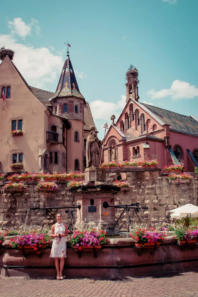 Caroline in front of the Saint-Leon fountain in Eguisheim, surrounded by flowering Alsatian houses