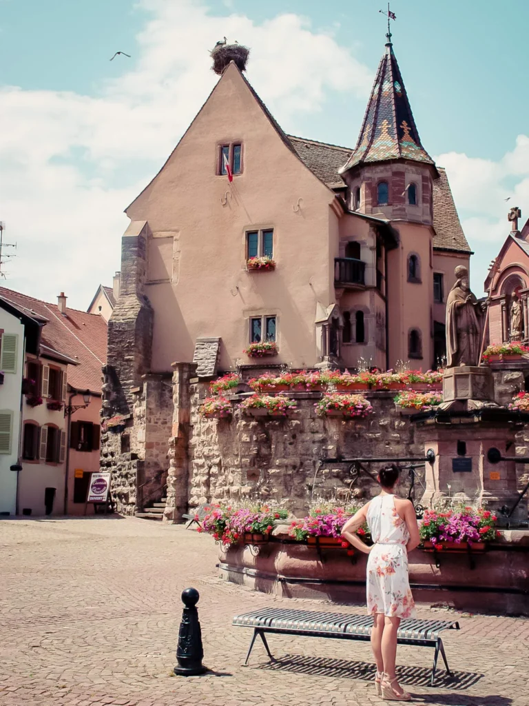 Caroline observing a flower-filled square and an Alsatian house in Eguisheim