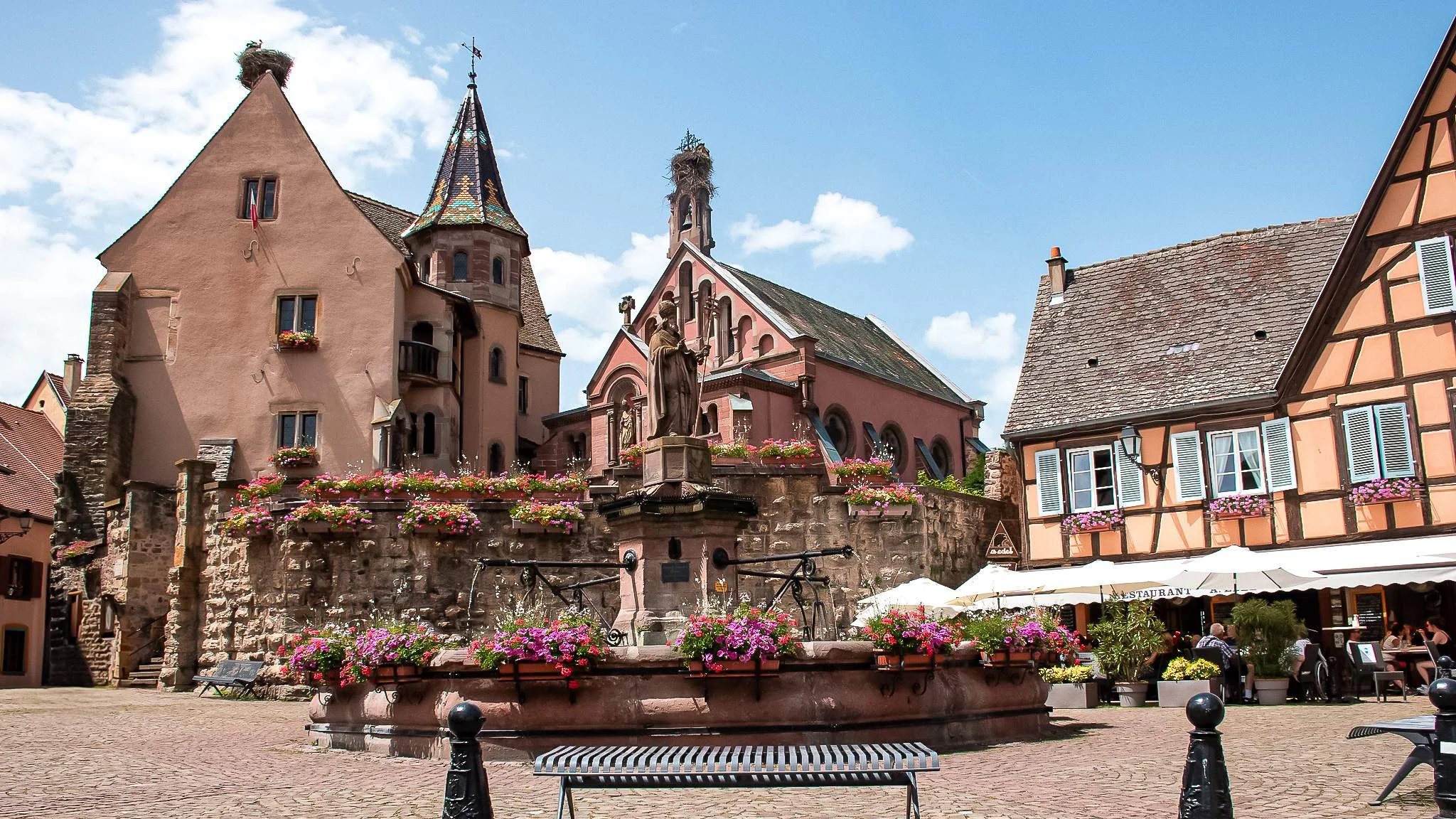 Place of Chateau Saint-Leon in Eguisheim: half-timbered houses, flowered fountain, and summer sky