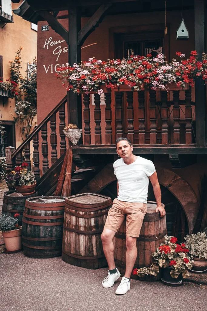 Xavier stands in front of a typical half-timbered house in Eguisheim, surrounded by wooden barrels.