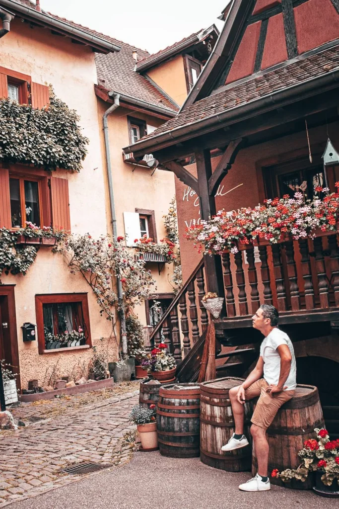Xavier sitting on a barrel in a picturesque alley of Eguisheim, surrounded by half-timbered houses