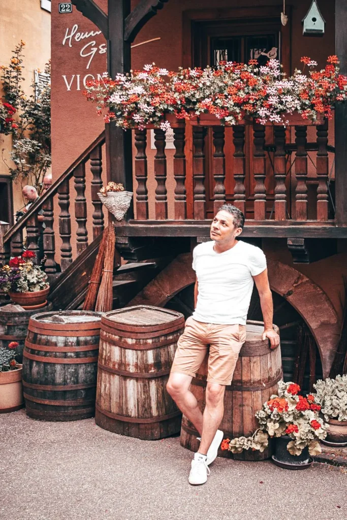 Xavier in front of a traditional Alsatian house in Eguisheim, surrounded by wooden barrels