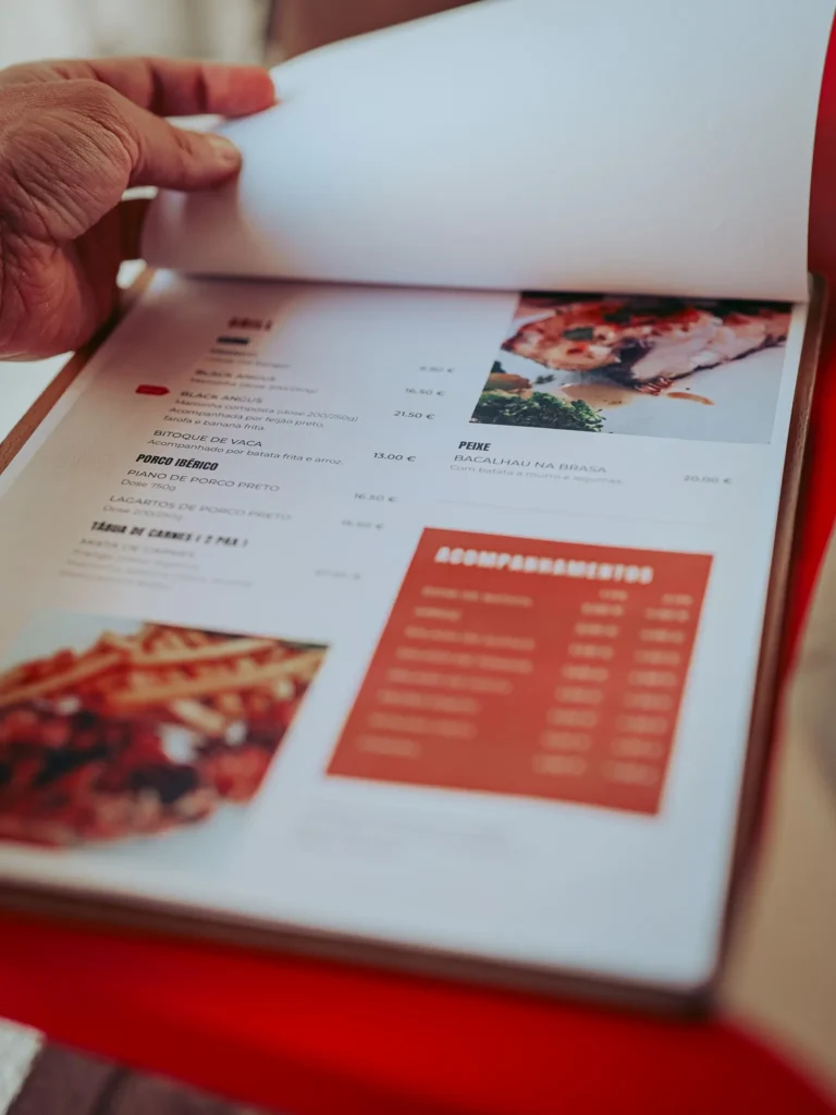 Menu of the Lumo restaurant in Tavira, Algarve, with a hand turning a page, showcasing typical dishes and sides.
