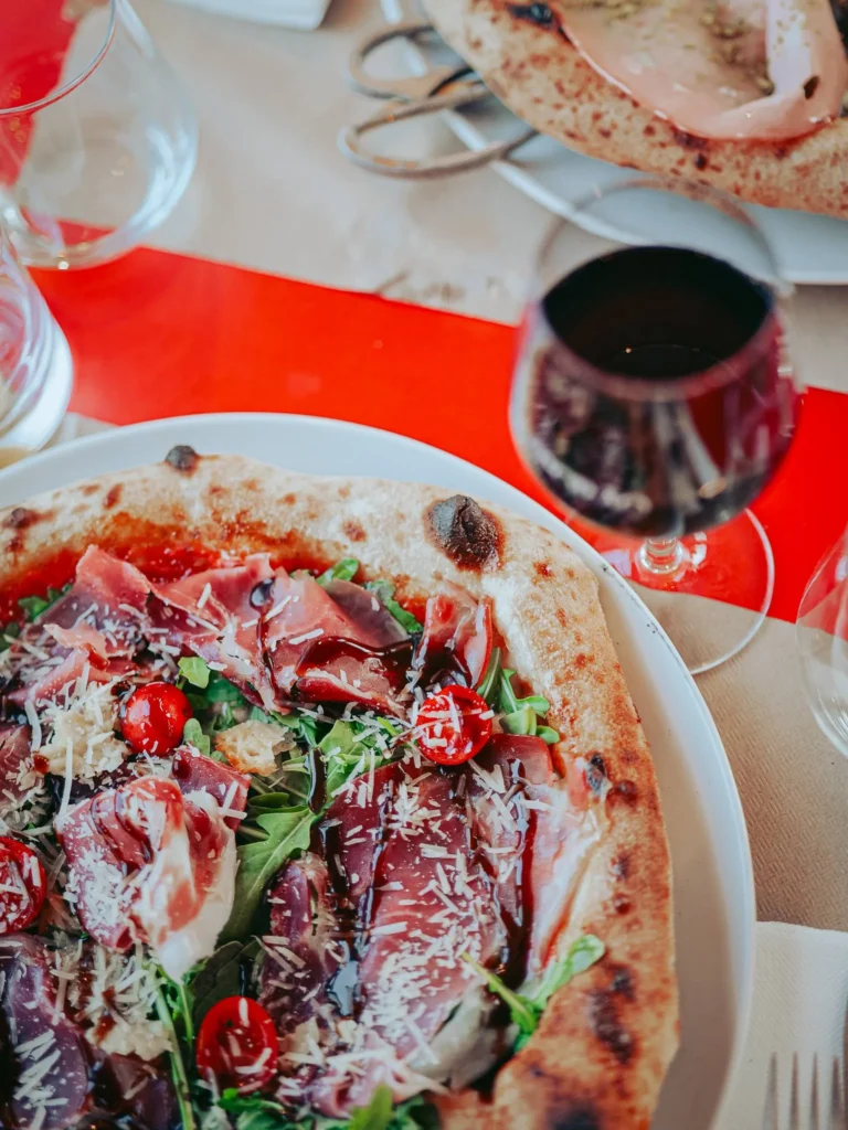 Pizza with prosciutto and arugula topped with Parma ham, rocket, cherry tomatoes, balsamic reduction, and pecorino, served on a beautifully set table at Lumo restaurant in Tavira, Algarve. A glass of red wine accompanies the dish.