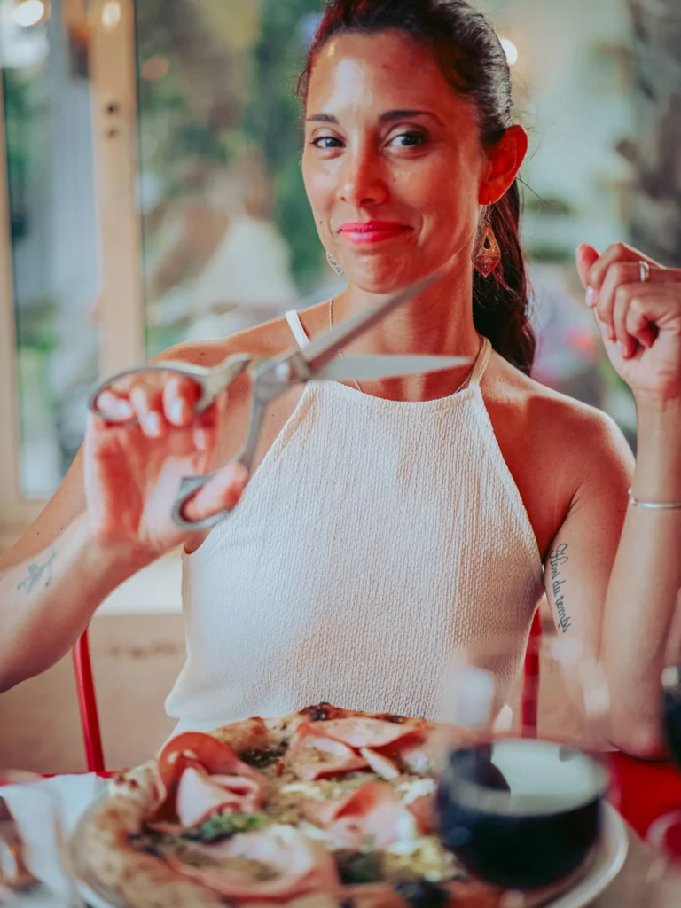 Caroline sitting at a table in the Lumo restaurant in Tavira, Algarve, cuts a pizza with scissors, a glass of red wine in front of her.