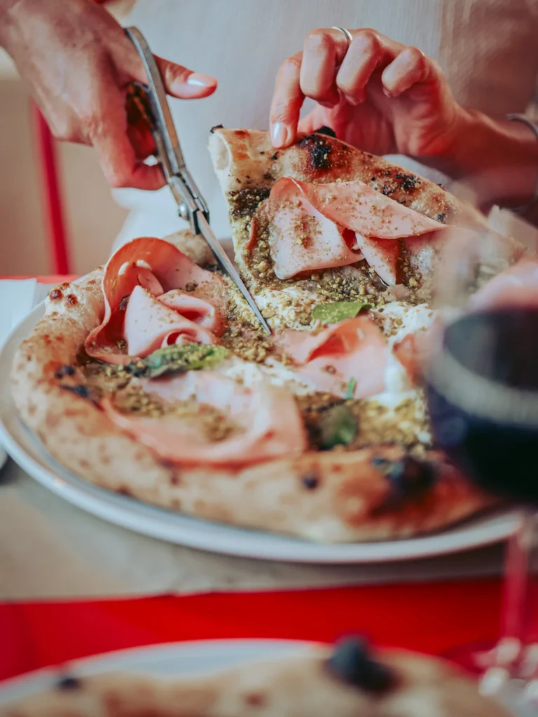 A pizza topped with mortadella, pesto, pistachio, and stracciatella is cut with scissors on a restaurant table in Tavira, Algarve.