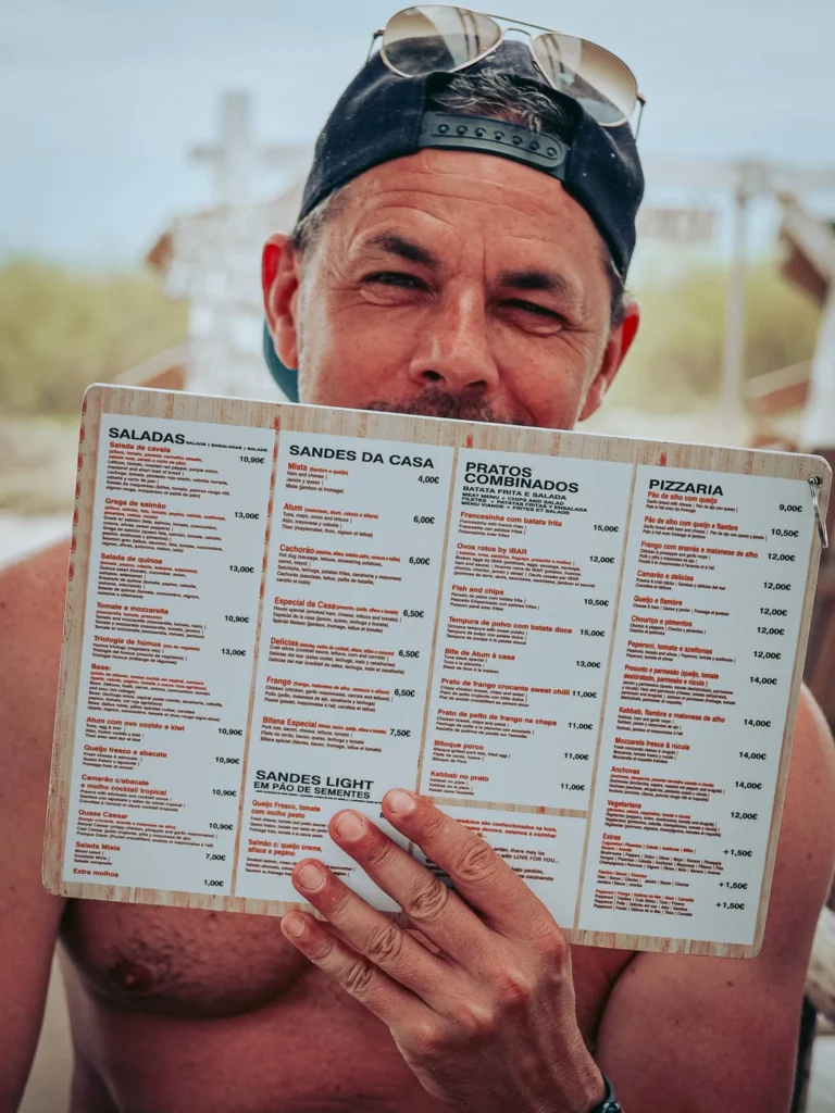 Xavier sitting on a terrace on the island of Tavira in the Algarve, holding a large restaurant menu in Portuguese in front of him. Summer and relaxed atmosphere.