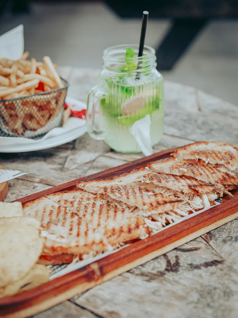 Close-up of a grilled chicken tosta served on a wooden board, accompanied by fries and a ginger and mint lemonade on a terrace table in Tavira, Algarve.