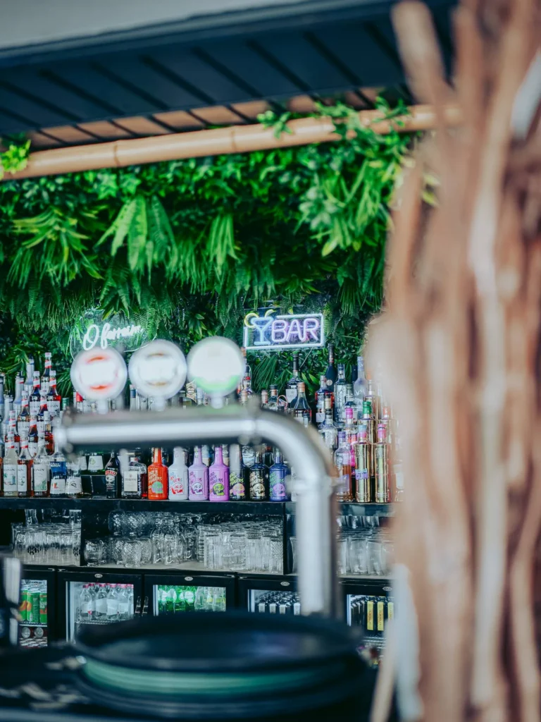 Outdoor bar counter at O Ferreira on Tavira Island, with numerous bottles of alcohol, glasses, and a neon sign, decorated with green plants.