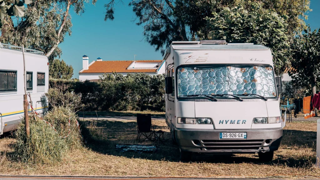 Hymer motorhome parked in a shaded spot at the Porto Covo campsite, with a white house and orange roof in the background and laundry hanging nearby.