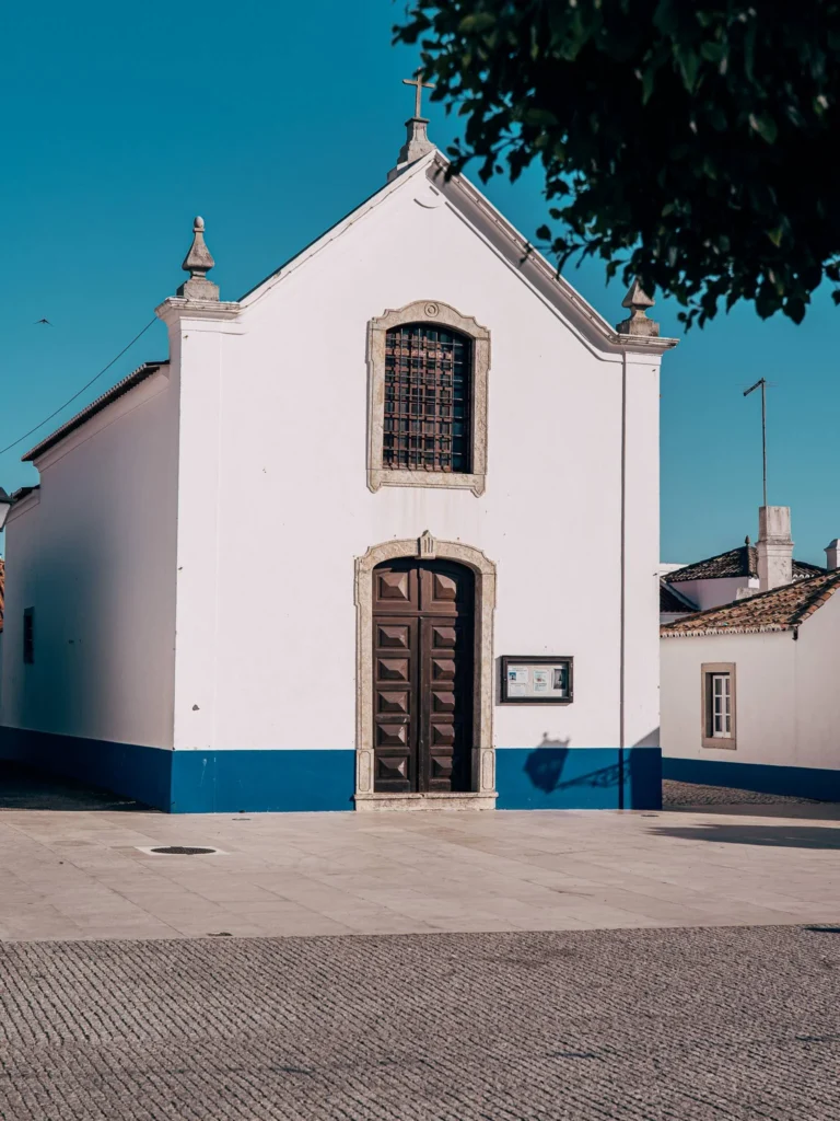 Small white chapel with blue details, typical of the architecture of Porto Covo in Alentejo, photographed under a clear sky in June.