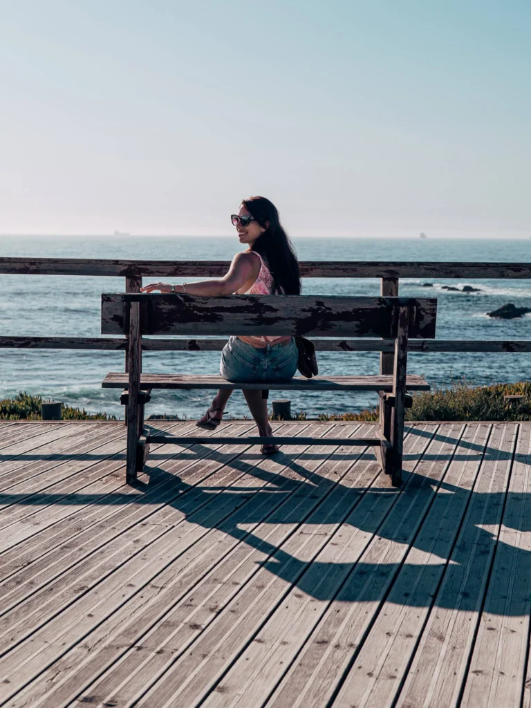 Caroline sitting on a wooden bench facing the Atlantic Ocean on a promenade in Porto Covo, under the early summer light.
