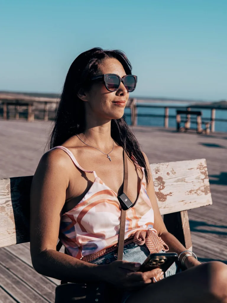 Caroline sitting on a wooden bench on a sunny pier in Porto Covo, enjoying the late afternoon light with the sea in the background.