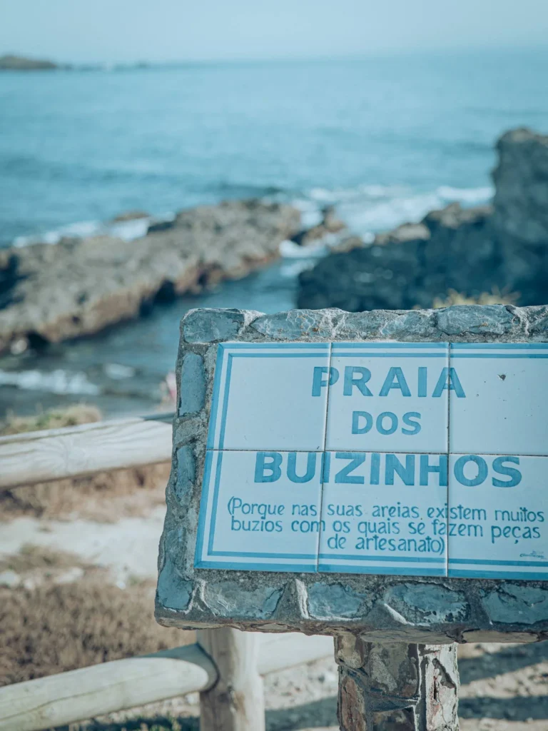 Ceramic sign indicating Praia dos Buizinhos in Porto Covo, with the rocky coast and the Atlantic Ocean under a clear sky in the background.