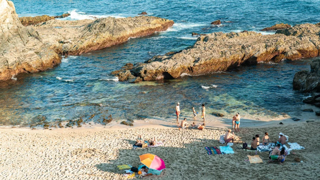 Golden sandy beach in Porto Covo, Alentejo, with several groups of people enjoying the sun and the sea near rocks by the Atlantic Ocean, in June.