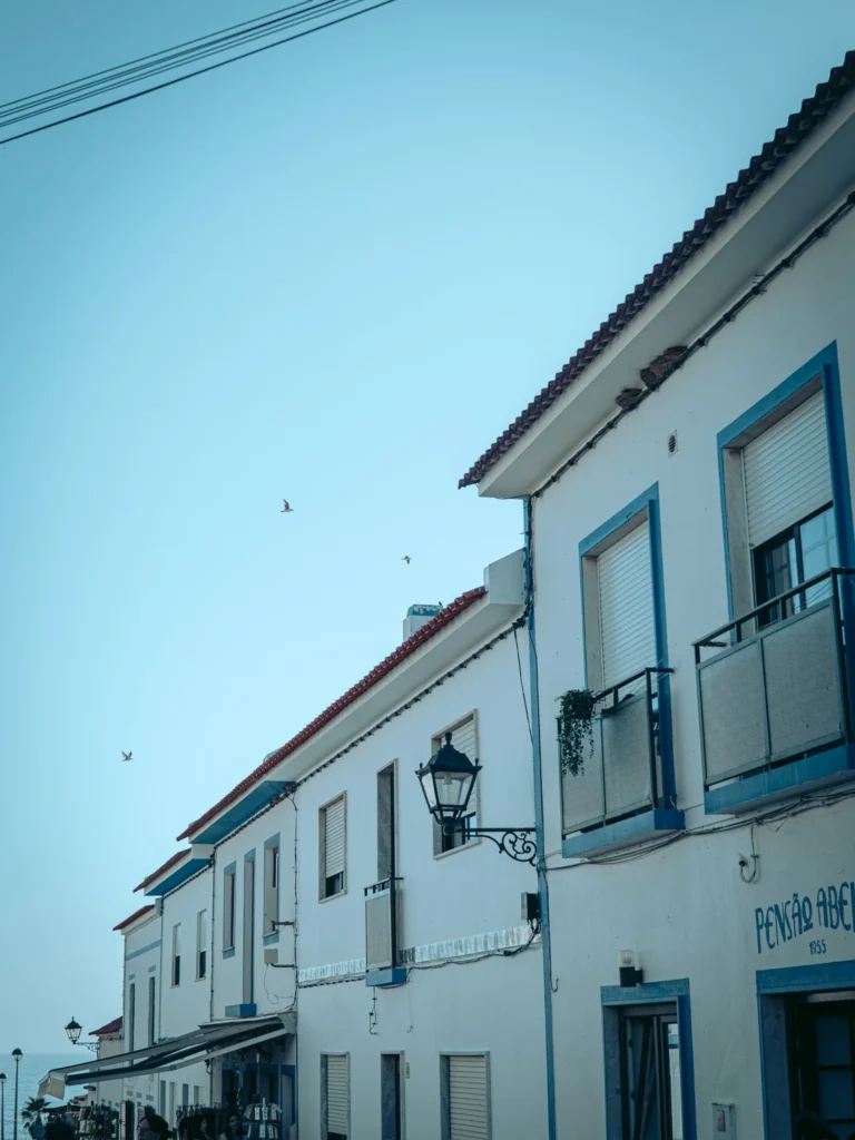 White house facades with blue shutters typical of Porto Covo, under a clear sky in Alentejo, Portugal.