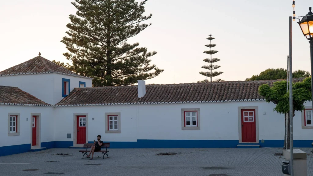 Xavier sitting on a bench in front of traditional white houses with red doors in Porto Covo, Alentejo, at the end of the day.
