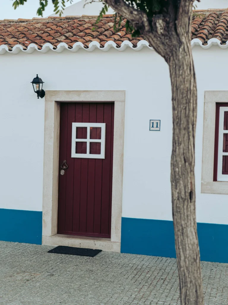 Facade of a traditional house in Porto Covo, with a burgundy wooden door, white walls, and a blue base, typical of Alentejo in Portugal.