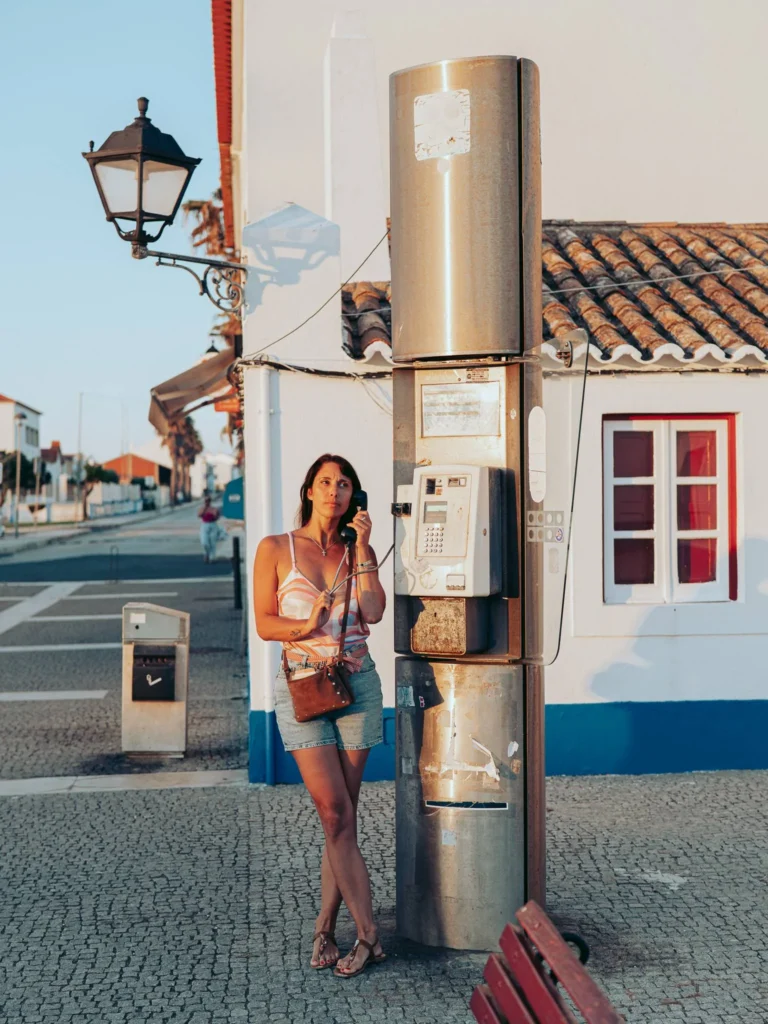 Caroline is using a phone booth on a sunny street in Porto Covo, with typical white houses of Alentejo in the background.