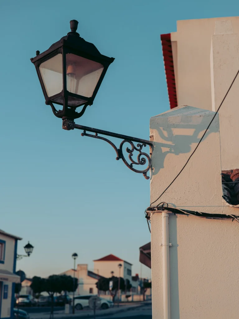 Antique wrought iron street lamp fixed to a white wall, in a quiet street of Porto Covo under the soft light of the late day.