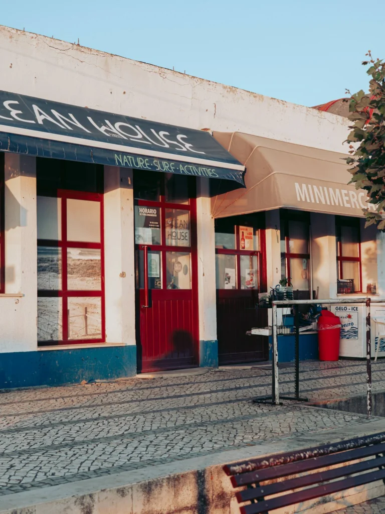 Facade of a small local shop in Porto Covo, Alentejo, with a minimarket sign and a sunny terrace in June 2026.